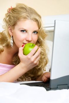 beautiful girl at home on the bed using a laptop and eating a green apple