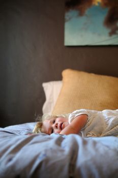 A blonde female toddler in a white dress relaxes playfully on her parents' bed.  The image is in selective focus.