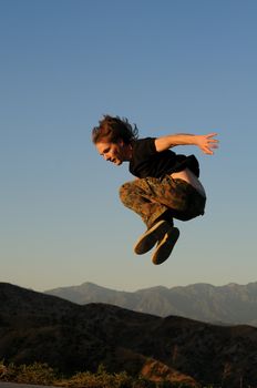 Caucasian male 20-40 in sportswear jumping above mountain peaks, face in profile to the left, one arm back, knees up.