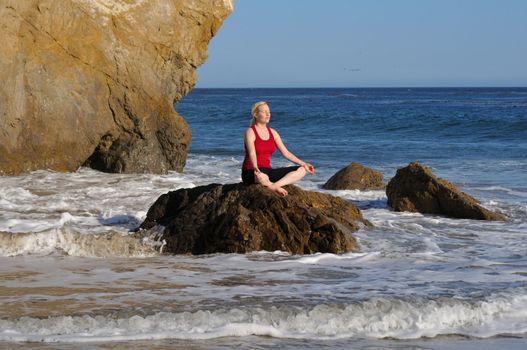 Woman 25-40 performs yoga meditation on a rock in the surf, facing the sun.