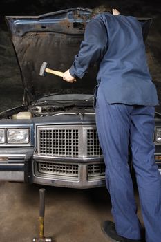 A bewildered mechanic about to use a hammer to take his frustrations out on the engine of an old car.
