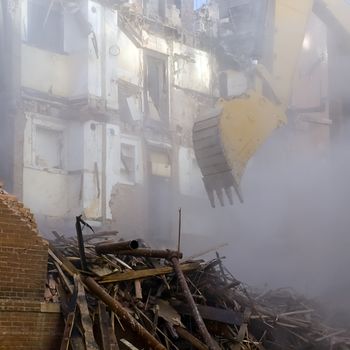 A backhoe emerging from the dust cloud to knock down the walls of a historic apartment building.