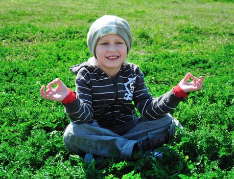 little boy is sitting on the green grass and doing meditation