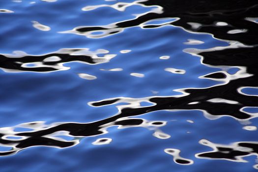 The shadow of a bridge reflects in the wavy water surface of a canal, creating a psychedelic kind of pattern. Cold colours: blue, black, white.
