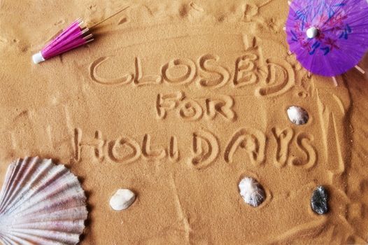Closed for holidays written on sand with colorful umbrellas and seashells. Soft focus for dreamy atmosphere.