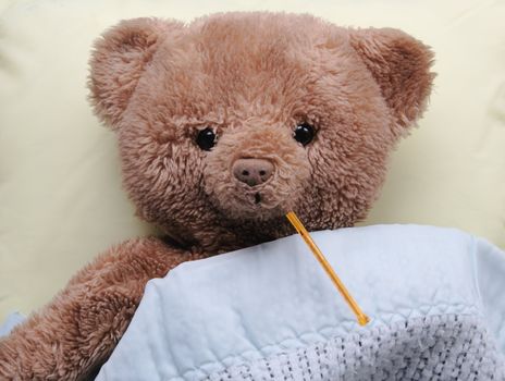 sad teddy bear on a pale yellow pillow with a thermometer, covered by a blue baby blanket
