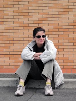 Casual girl with sunglasses sitting in front of a brick wall.          