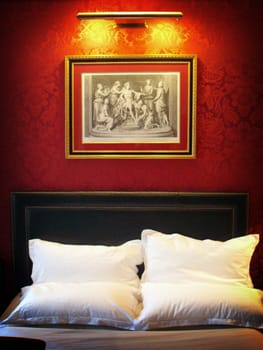 Red wallpaper with art hanging on the wall above the top end of a bed in a Paris Hotel room.