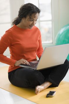 Beautiful young African American woman sitting on yoga mat using her laptop