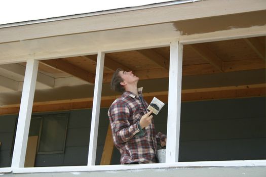 Boy with bucket of white paint looking up as he paints porch on house