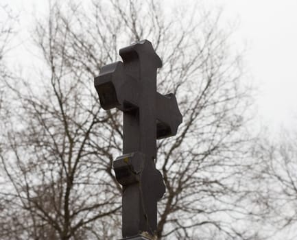 Black marble orthodoxal cross-shaped tombstone against intertwined tree branches on a gloomy winter day