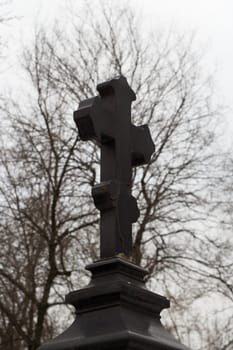 Black marble orthodoxal cross-shaped tombstone against intertwined tree branches on a gloomy winter day