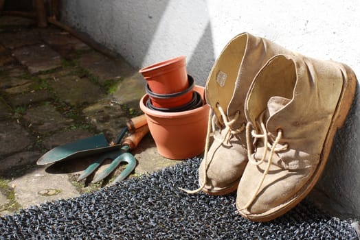 A pair of gardening boots resting against a wall with terracotta coloured pots and garden tools.