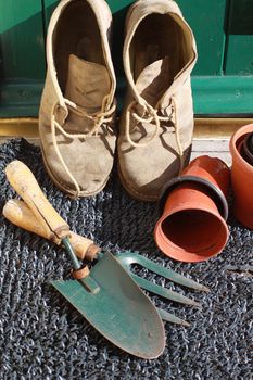 A pair of gardening boots resting against a green wooden back door with terracotta coloured pots and gardening tools surrounding.