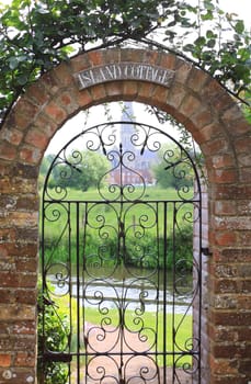 View looking through a country cottage garden gate with a view of Salisbury Cathedral in distance. Gate surrounded by a brick constructed arch covered with a rambelling rose plant.