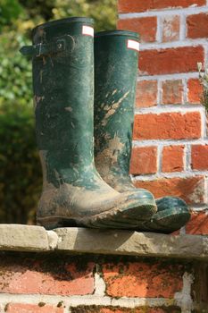 A pair of gardening boots set on a patio step. Backdrop consists of the end of a bricked building.
