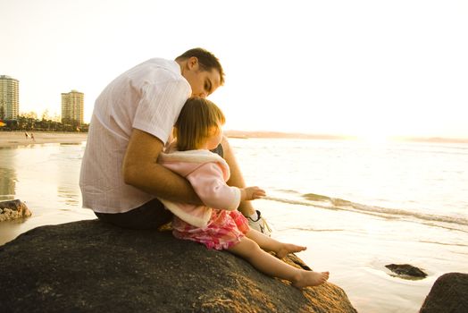 Little girl and her dad sitting on a rock at a beach watching the sun go down