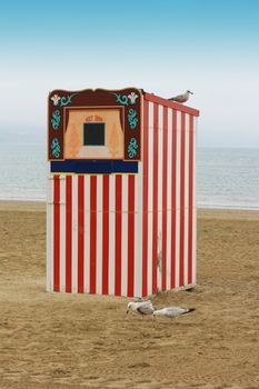 A red and white striped wooden punch and Judy booth on a beach located in Weymouth, Dorset UK. 
A seagull perches on top, with two seagulls pecking at the sand below.