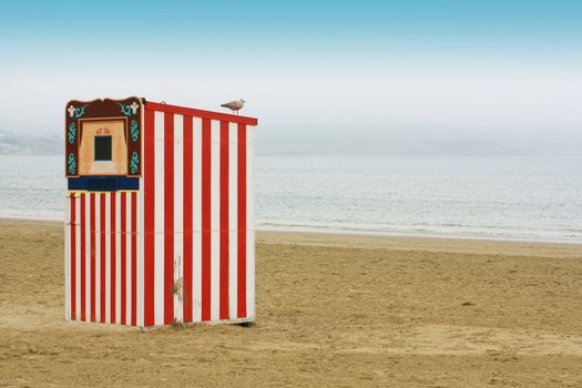 A red and white striped wooden punch and Judy booth on a beach located in Weymouth, Dorset UK. 
A seagull perches on top. landscape format, room for copy space etc.