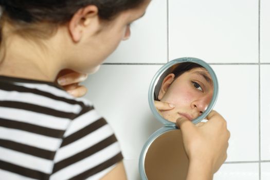 Young teenage female holding a mirror looking at her complexion with concern.
