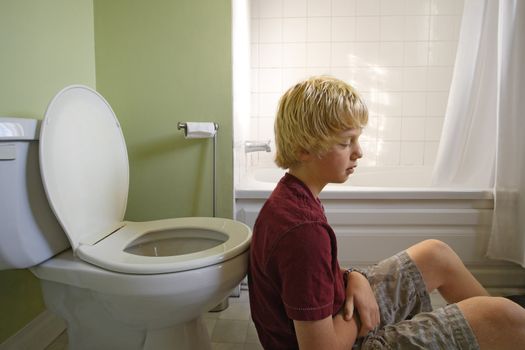 A young boy suffering with a stomachache, leaning up against a toilet. Shot with natural light coming through a window.

