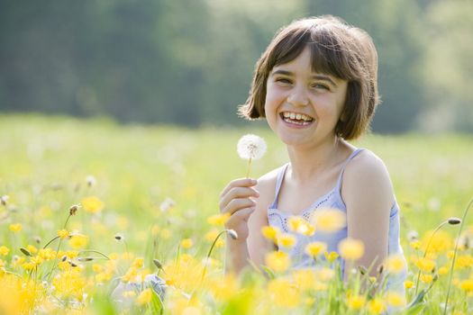 young female child sitting cross legged in a field full of buttercups holding dandelion and smiling at camera with room for copy