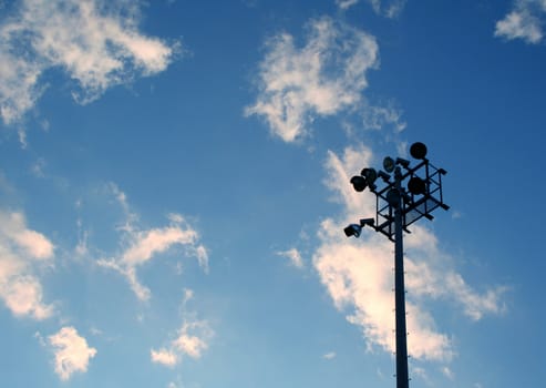 The silhouette of stadium light stand set against a blue sky with white clouds.
