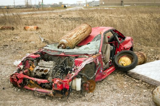 This is an abandoned red sports car in a field near a factory.