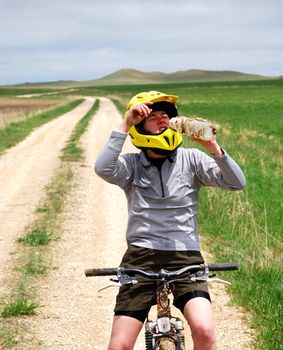 Mud-splattered mountain biker drinking water against a dirt road and green pasture.