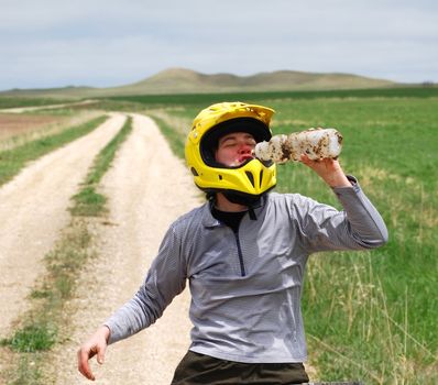 Mud-splattered mountain biker drinking water against a dirt road and green pasture.