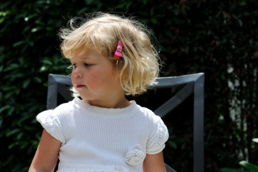 Little girl sitting outside in a garden on a chair, gazing pensively off to the side.