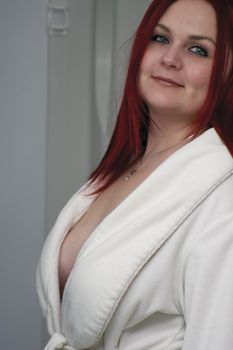 Red hair woman model in open white bathrobe standing in bathroom