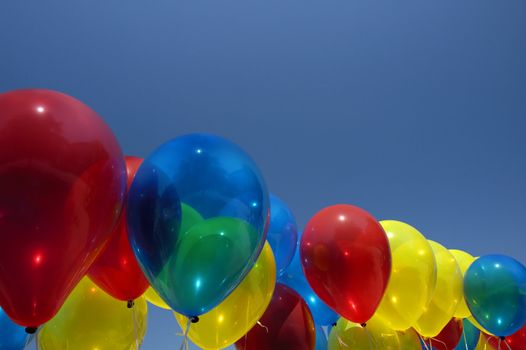 A row of multi-coloured balloons floating against a clear blue sky. A tree can be seen, reflected in the blue one. Space for text in the sky.
