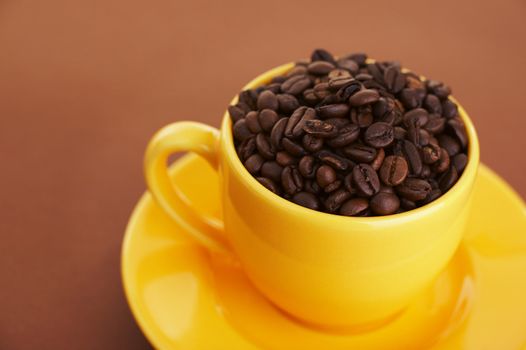 Yellow coffee cup filled with coffee beans over a brown background (shallow dof)