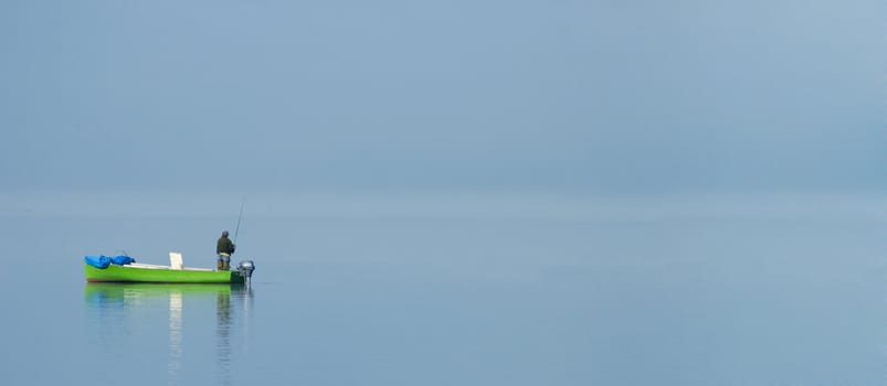 A fisherman stands alone in his boat on a misty lake, shortly after dawn. Space for text.