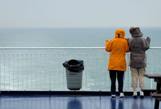 Two young women, in the rain, looking over the rail at a grey sea and waving. A container ship can just be seen on the horizon. Maybe they know someone on the ship.