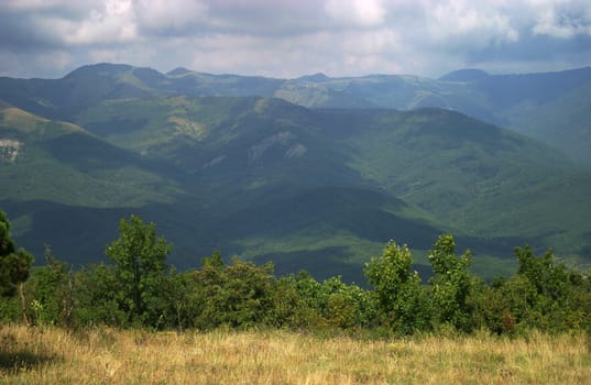 The image of mountains with shadows from clouds and a glade with a dry grass and green bushes