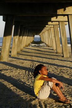 An African-American girl sitting on the beach with a pier in the background.