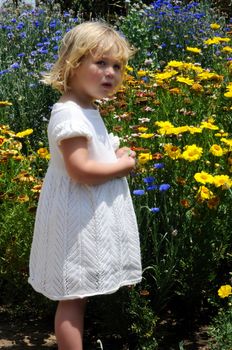 Little girl stands in front of a profusion of blue and yellow flowers, holding a leaf in her hands.