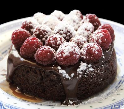 an individual-sized chocolate cake topped with ganache, fresh raspberries, powdered sugar and caramel sauce