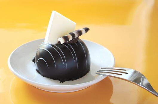 a chocolate dessert with a white chocolate decoration on a white plate with a fork against a reflective medium yellow background
