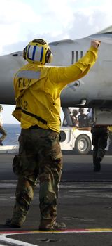 A Navy sailor directs an F-18 fighter aircraft around the flight deck on an aircraft carrier