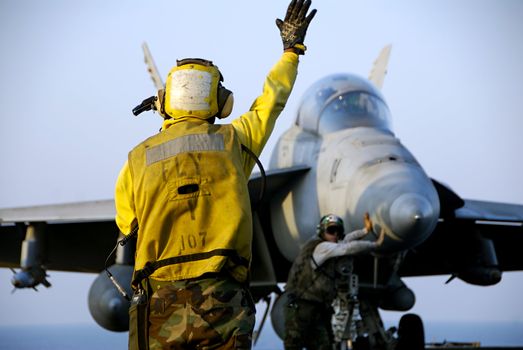 A Navy Sailor directs an F-18 Hornet fighter aircraft around the flight deck of an aircraft carrier