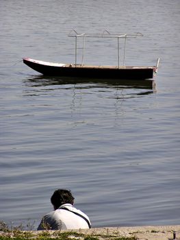 Man sitting at river bank with calm river and boat in background.