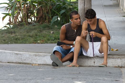 Two guys sitting on sidewalk and looking at something. January 2008, Camaguey, Cuba.