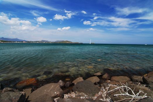 Beautiful sea scenery on Sardinia - stones in the front, sail and blue sky with clouds