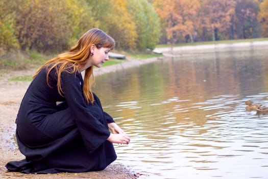 Girl in black dress sitting near pond with ducks
