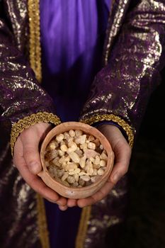 Wise man arrayed in purple cloak embrodered with metallic gold thread, his hands holding an ancient clay pot filled with the best hojari frankincense resin tears from dhofar region of Oman.  The aroma is warm and citrusy