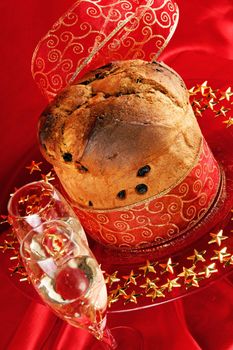 Panettone the italian Christmas fruit cake with red and golden ribbon served on a red glass plate over a red background. Two glasses of champagne in the foreground. Selective focus.