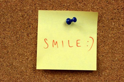Yellow small sticky note on an office cork bulletin board. Smile!